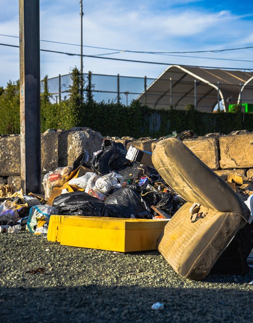 A pile of mixed waste and rubbish, including black plastic bags, a broken beige car seat, and a yellow plastic container, is shown on a gravel surface beside a small stone wall. The rubbish appears to