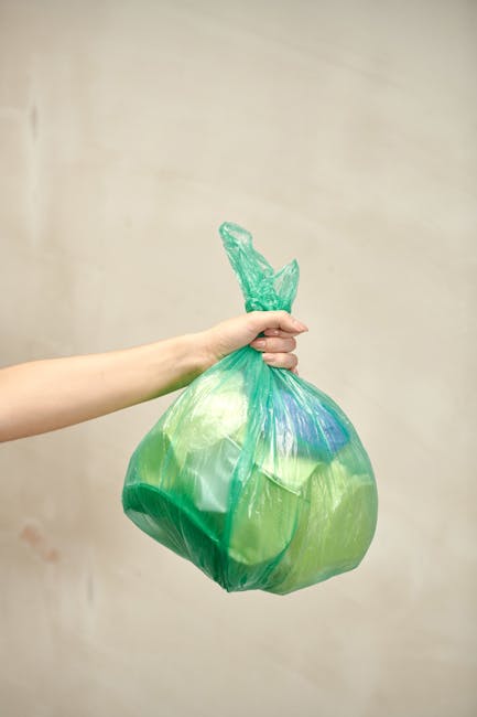 A pile of mixed waste and rubbish, including black plastic bags, a broken beige car seat, and a yellow plastic container, is shown on a gravel surface beside a small stone wall. The rubbish appears to